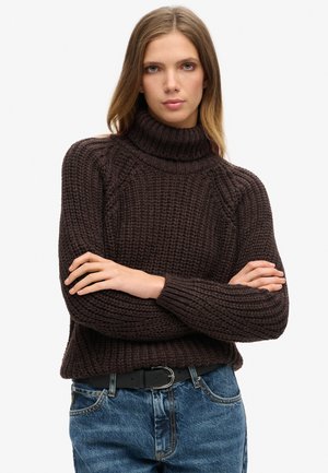 Brun strikket turtleneck sweater med ribbede manchetter og kant, parret med blå denim jeans og et sort bælte med en metalspænde.