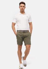 Polo shirt blanc à manches courtes avec un logo, associé à un short vert olive ; il présente un ourlet roulé et une ceinture à rayures multicolores. Des baskets complètent le look.