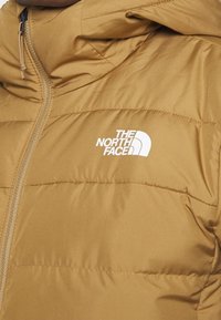Braune Daunenjacke mit Kapuze, die über gesteppte Abschnitte und ein weißes Logo verfügt. Der Stoff sieht glatt aus und hat eine glänzende Oberfläche sowie einen Reißverschluss.