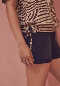 Korte mouwen, linnen top met bruine strepen, gecombineerd met donkere denim shorts met opgerolde zomen en een zijstrikdetail. Gouden ring aan de hand.