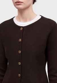 Personne portant un cardigan marron foncé boutonné sur une chemise blanche, montrant des boutons dorés texturés et un col rond.
