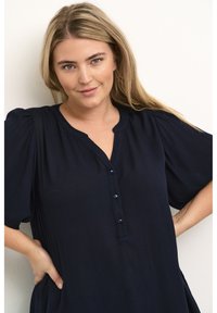Camicia tunica blu navy con maniche corte a sbuffo, scollatura a V e dettagli con bottoni sul davanti. Realizzata in un tessuto leggero e testurizzato.