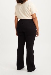 Jean flare noir à taille haute, associé à un t-shirt rose clair. Poches arrière visibles et texture en coton lisse. Baskets blanches.