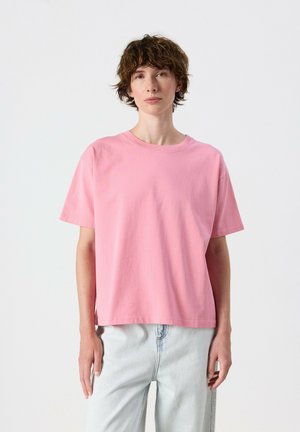 Personne avec des cheveux courts et bouclés portant un t-shirt rose ample et un jean bleu clair, debout devant un fond blanc uni.