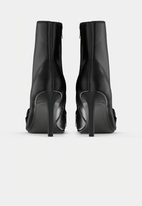 Bottines noires à talons hauts avec fermeture éclair au dos et talons aiguilles pointus, vues de dos sur un fond uni.