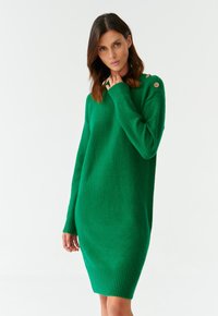 Robe pull en tricot vert avec des manches longues, des poignets et un ourlet côtelés, et des boutons décoratifs sur l'épaule, présentant un tissu doux et texturé.