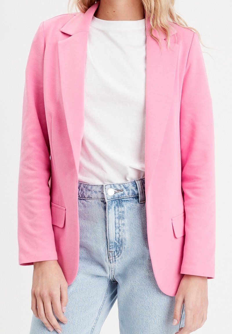 Blazer en tissu extensible rose avec revers à crans, poches latérales et coupe cintrée, porté sur un t-shirt blanc et un jean bleu taille haute.