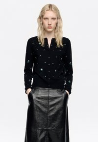 Melnā jaka ar pogām, ar zaļiem un sudraba rotājumiem. Valkāta virs spīdīgas melnas ādas svārku. Gluda tekstūra un pieguļošs dizains.