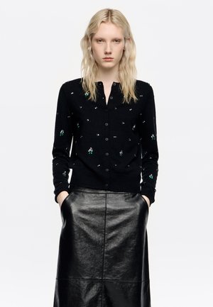 Czarny kardigan z guzikami, zdobiony zielonymi i srebrnymi detalami. Nosi się go na błyszczącej czarnej skórzanej spódnicy. Gładka faktura i dopasowany krój.