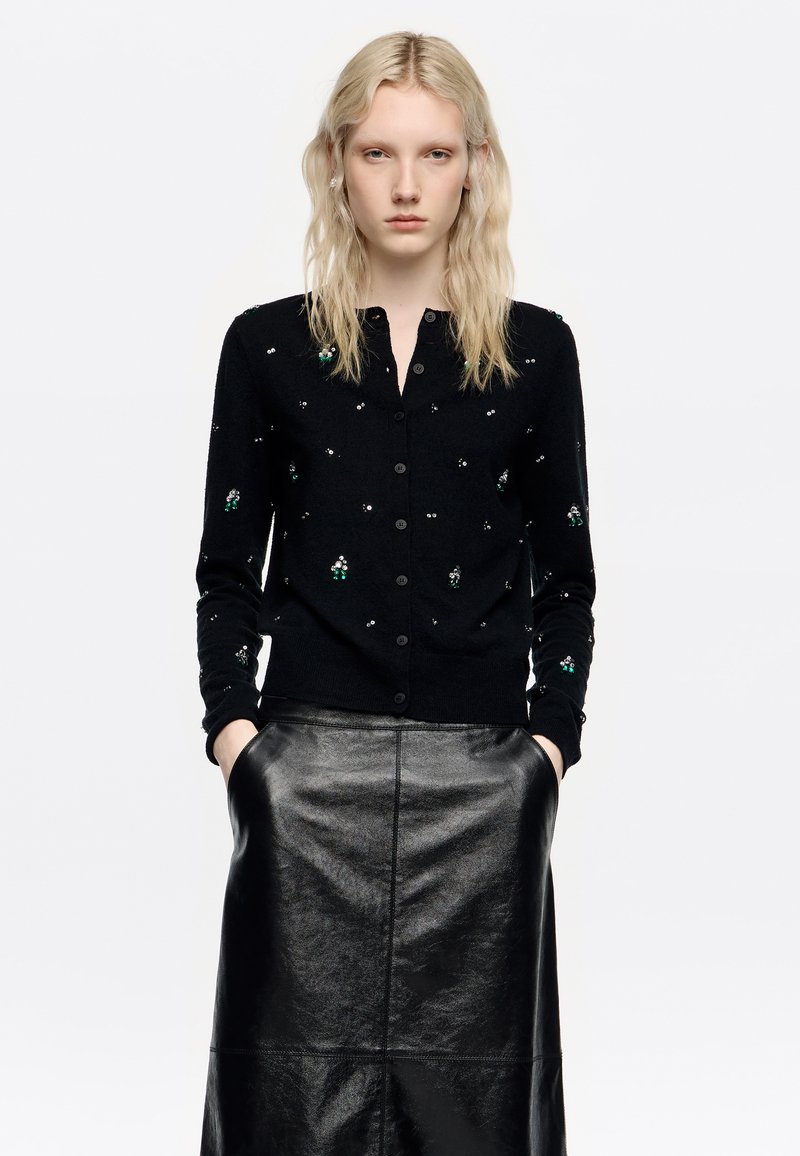 Melnā jaka ar pogām, ar zaļiem un sudraba rotājumiem. Valkāta virs spīdīgas melnas ādas svārku. Gluda tekstūra un pieguļošs dizains.