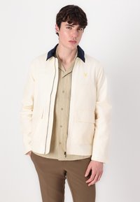 Cream-colored jacket with a navy collar, front zip, and two patch pockets. Worn over a light green, short-sleeved, ribbed shirt.