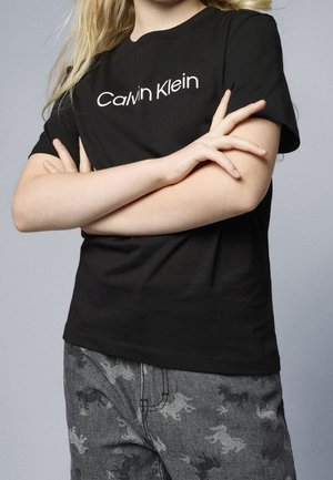 Enfant bras croisés portant un t-shirt Calvin Klein noir et un pantalon gris avec des motifs animaux sombres sur un fond uni.