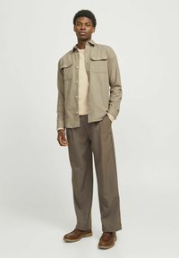Chemise kaki à boutons avec deux poches poitrine, portée par-dessus un pull beige et associée à un pantalon brun ample et des chaussures brunes.