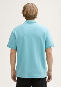 Lichtblauwe polo met korte mouwen van een gestructureerde stof, standaard kraag en zijnaadlabeldetail, gedragen met donkere broeken.