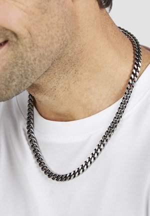 Massive silberne Halskette mit abwechselnd polierten und matten Gliedern, die auf einem weißen Hemd liegt und eine glatte, reflektierende Oberfläche aufweist.