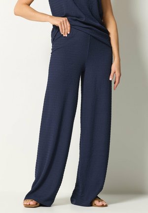 Frau trägt marineblaue, strukturierte, weit geschnittene Hose mit passendem Oberteil, steht mit einer Hand in der Hüfte und sichtbaren Sandalen.