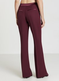 Donna che indossa pantaloni svasati a vita alta di un classico bordeaux scuro, con tasche posteriori a filetto, in piedi a piedi nudi su uno sfondo chiaro e semplice.