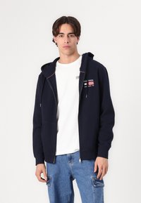Sweat à capuche bleu marine avec fermeture éclair, doté d'une capuche à cordon, portant un patch logo et des accents rouges, porté sur un t-shirt blanc et associé à un jean bleu clair.
