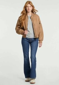 Καφέ bomber jacket με ριγέ μανίκια, γκρι μπλούζα, φαρδιά μπλε τζιν και καφέ αθλητικά παπούτσια. Το μοντέλο στέκεται μπροστά σε ουδέτερο φόντο.