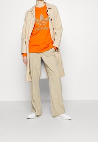 Abrigo trench beige sobre un sudadera naranja con un logo plateado, combinado con pantalones de pierna ancha beige a rayas y zapatillas blancas.