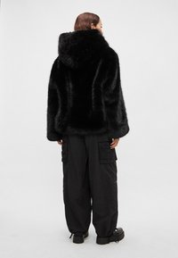 Juoda dirbtinio kailio striukė su gaubtu, pūkuota faktūra, plačiais rankogaliais ir laisvo silueto, derinama su juodomis karo stiliaus kelnėmis ir masyviais batais.