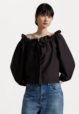 Blusa preta off-shoulder com decote franzido e mangas bufantes, feita de tecido texturizado, combinada com jeans de denim azul claro.