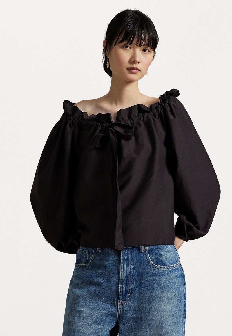 Black off-shoulder blouse with ruffled neckline and puffed sleeves, made from textured fabric, paired with light blue denim jeans.