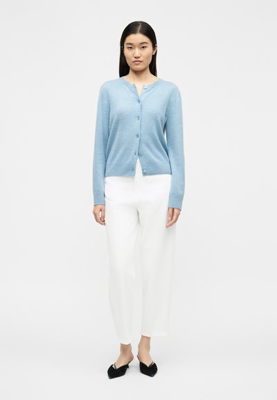 Γυναίκα που στέκεται φορώντας μια ανοιχτό μπλε ζακέτα με κουμπιά, λευκό παντελόνι ψηλόμεσο και μαύρα μυτερά παπούτσια με τακούνι σε λευκό φόντο.