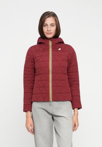 Veste matelassée rouge avec une capuche, dotée d'une fermeture éclair à l'avant avec un accent jaune, de motifs en losange et d'un logo sur le côté gauche.