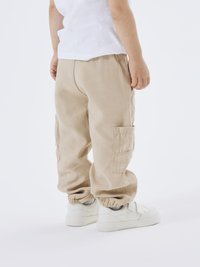 Pantalones cargo beige con múltiples bolsillos laterales, puños elásticos y un ajuste relajado. Llevados con zapatillas blancas. Vista trasera.