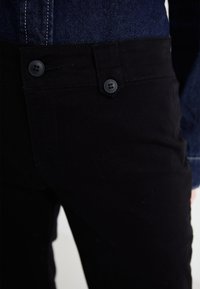 Black cotton pants featuring a flat front design, two black buttons, and contrasting stitching, paired with a denim top.