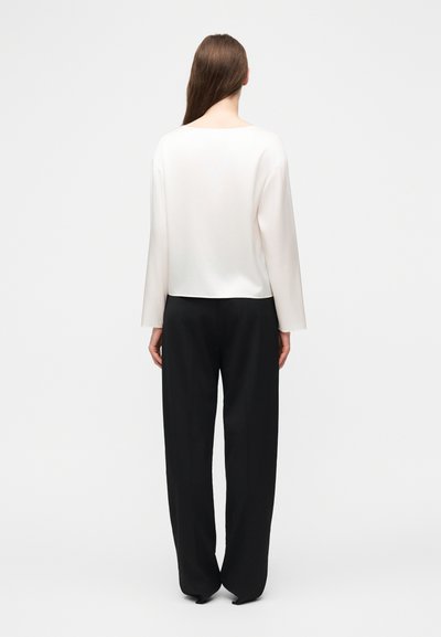 Γυναίκα με μακριά καστανά μαλλιά που κοιτάζει προς τα πίσω, φοράει λευκό μακρυμάνικο πουλόβερ και φαρδιά μαύρα παντελόνια μπροστά σε απλό ανοιχτόχρωμο φόντο.