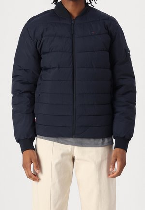 Mies pukeutuneena laivastonsiniseen tikattuun bomber-takkiin, jossa on vetoketju, resorit kädenteissä sekä pienet merkkilogot rinnassa, hihassa ja helmassa.