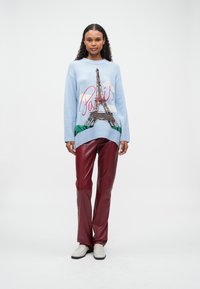 Frau in einem hellblauen Pullover mit dem Eiffelturm und dem Schriftzug "Paris", kombiniert mit roten Lederhosen und weißen Schuhen, stehend vor einem weißen Hintergrund.