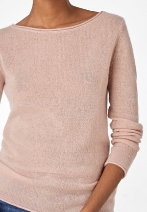 Femme portant un pull doux, rose clair, à manches longues avec un large décolleté et des manches légèrement relevées.