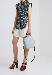Camicia corta a maniche corte verde scuro con motivo floreale; pantaloni corti color crema; sandali con tacco marroni; borsa azzurro chiaro con dettagli dorati e tracolla.