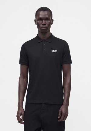 Homme portant un polo noir Karl Lagerfeld à manches courtes, debout devant un fond clair uni.