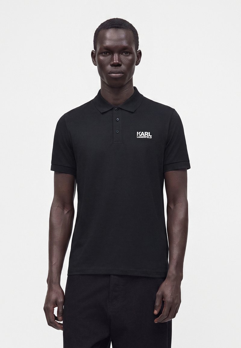 Homme portant un polo noir Karl Lagerfeld à manches courtes, debout devant un fond clair uni.