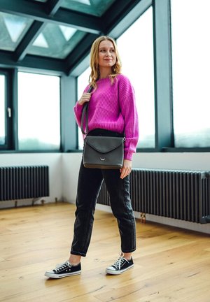 Black crossbody bag with a curved flap, worn with a bright pink knitted sweater and black denim pants. Black low-top sneakers. Wood floor.