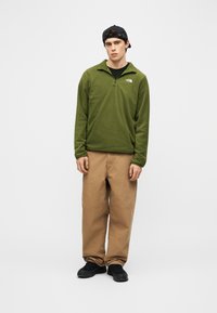 Νέος άνδρας που φοράει μαύρο καπέλο, φλις με μισό φερμουάρ σε ελαιοπράσινο χρώμα, μπεζ παντελόνι και μαύρα παπούτσια, στέκεται μπροστά σε απλό ανοιχτόφωτο φόντο.