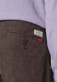 Gros plan d'une poche de pantalon marron Levi's avec bouton et étiquettes de la marque portée avec un haut violet clair.