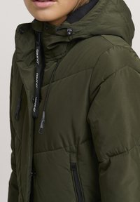 Oliven grønn dunjakke med hette, quiltet tekstur, justerbare snorer og sidelommer med glidelås. Stoffet ser ut til å være vannavstøtende.