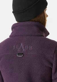 Lila Fleecejacke mit hohem Kragen, mit einem Logo "SLADE" auf dem Rücken und einem Metallhaken-Detail am Hals.