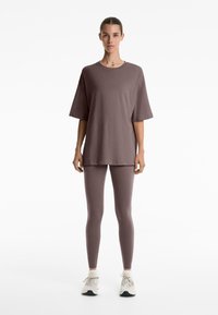 Svetlohnedé oversized tričko a zodpovedajúce legíny z mäkkej, elastickej tkaniny. Model má na sebe biele tenisky a stojí proti jednoduchému pozadiu.