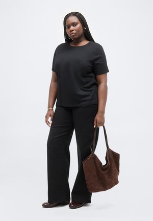 Žena s dlhými zapletenými vlasmi, oblečená v čiernej košeli a nohaviciach, drží veľkú hnedú semišovú kabelku na jednoduchom pozadí.