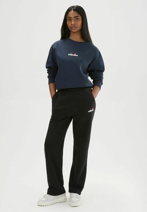 Frau steht mit den Händen in den Taschen, trägt einen marineblauen Sweatshirt und schwarze Hose, beide mit „ellesse“-Logo, und weiße Turnschuhe.