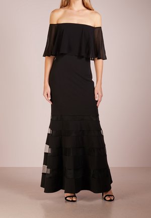 Kvinde iført en lang sort off-shoulder kjole med lagdelte transparente flæser i bunden, stående mod en ensfarvet baggrund.