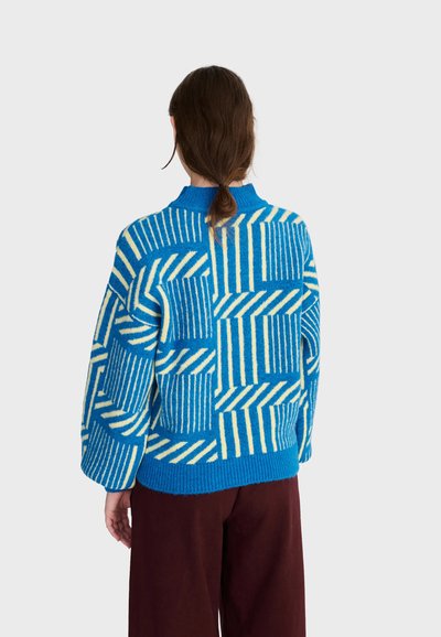 Personne aux cheveux bruns attachés bas portant un pull bleu et blanc à motifs et un pantalon marron foncé, de dos sur un fond uni.