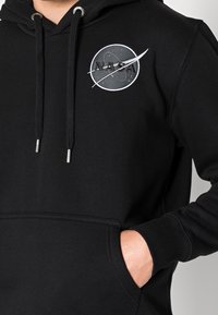 Persona que lleva una sudadera con capucha negra con el logo de NASA en blanco y negro en el pecho, con la mano en el bolsillo delantero y los cordones negros colgando.