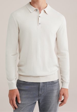 Mann trägt einen cremefarbenen Poloshirt-Pullover mit langen Ärmeln, kombiniert mit ausgewaschenen grauen Jeans und steht vor einem schlichten Hintergrund.